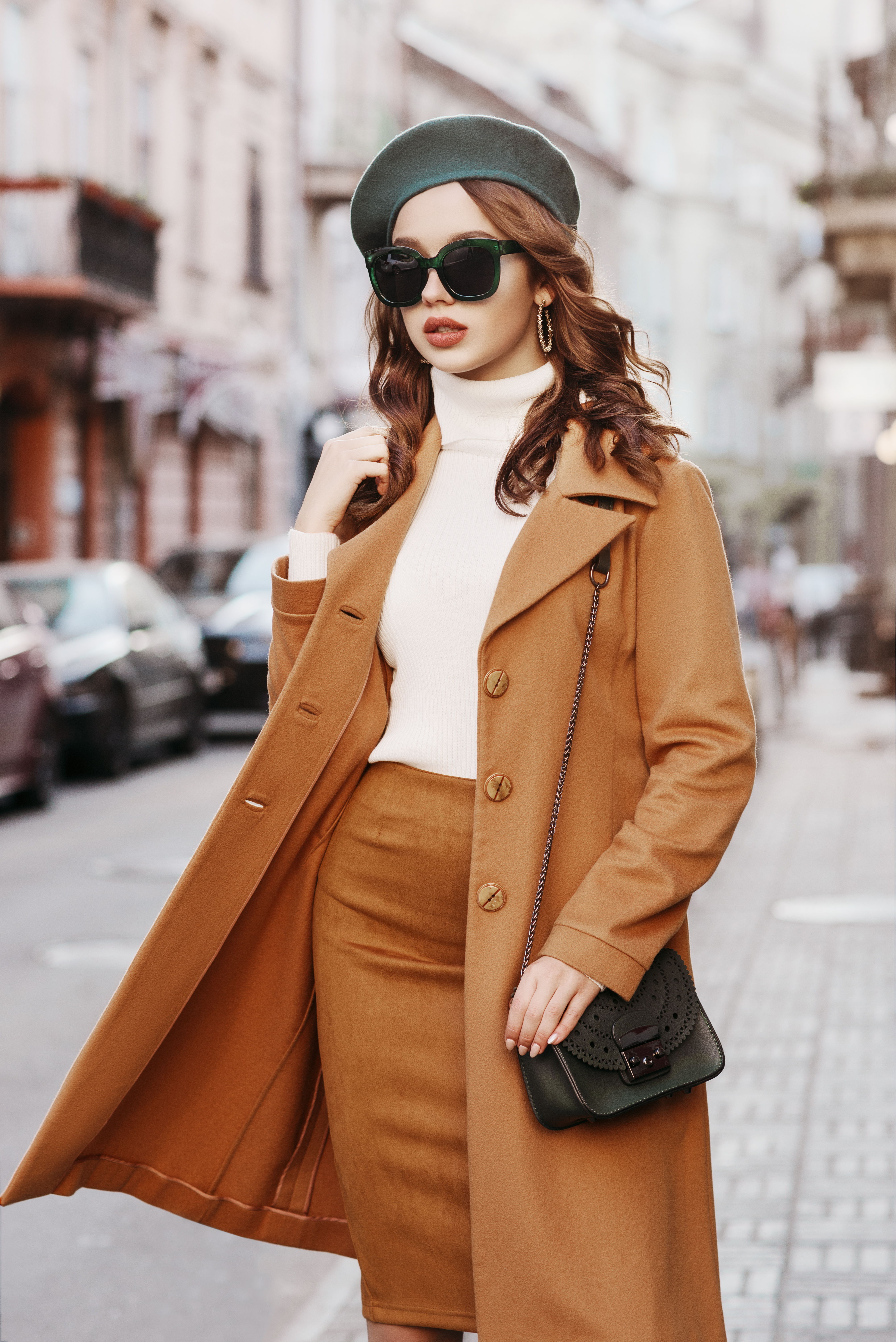 Outdoor portrait of young beautiful fashionable woman wearing trendy autumn coat, turtleneck, skirt, beret, sunglasses, with small shoulder bag, walking in street of european city