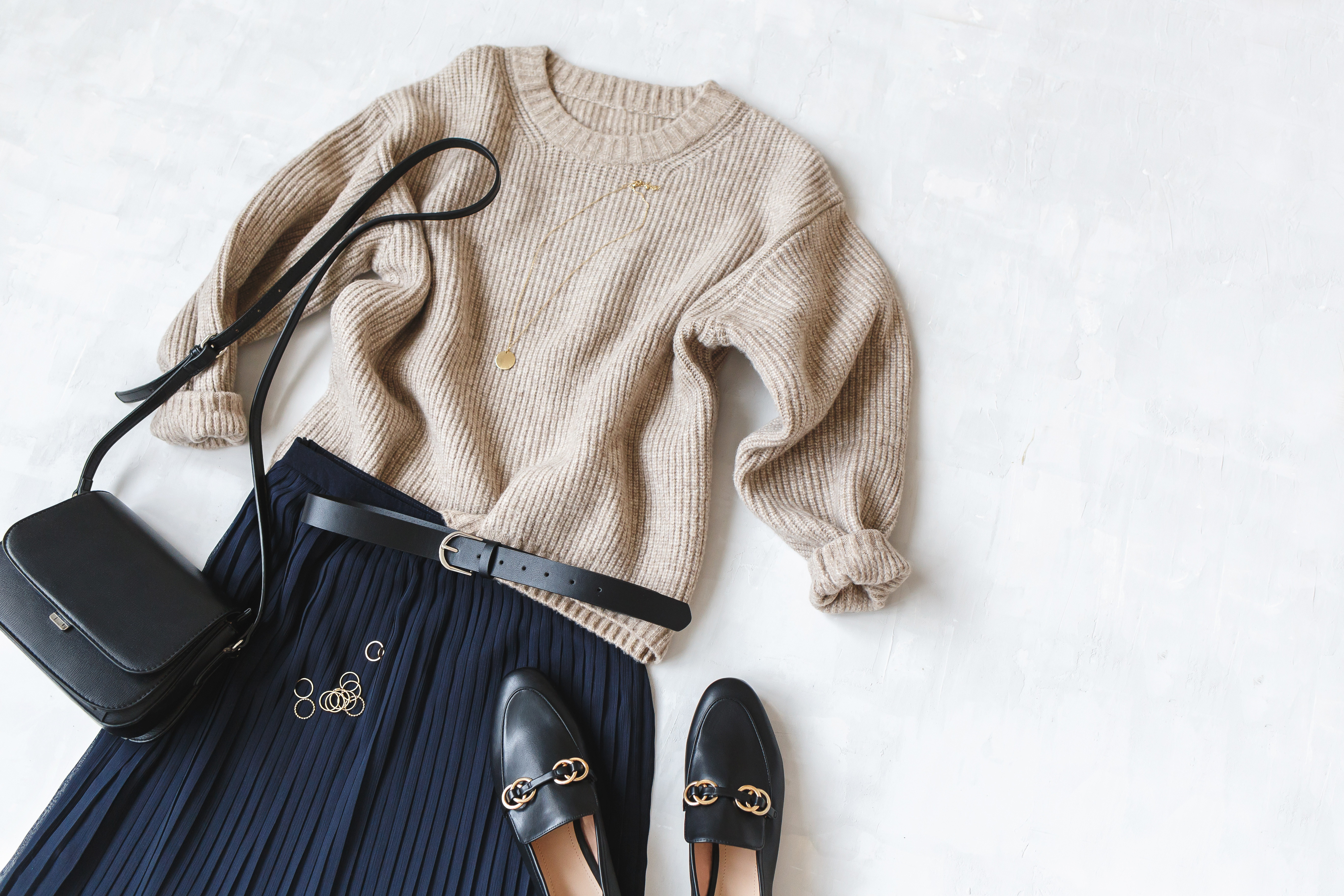 Blue midi pleated skirt, beige knitted sweater, small black cross body bag, belt, loafers (flat shoes) on grey background. Overhead view of women's casual day outfit. Flat lay, top view. Women clothes